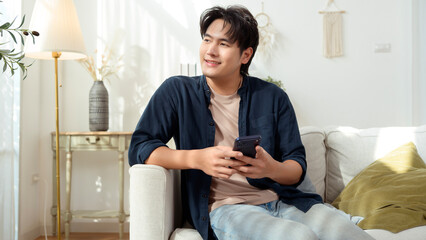 Obraz premium Young Man Using Smartphone While Sitting on Couch in Modern Living Room During Daytime