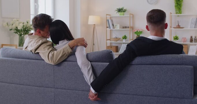 Back view of three on a sofa the suited rival holds her hand, implying cheating, infidelity, affair, triangle, betrayal, deception in the couple. Secret handholding betrays a hidden affair.