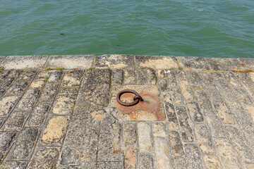 Fototapeta premium A weathered iron mooring ring is set into an aged stone quay by green harbor water in Locmariaquer. Sunlit masonry displays rough textures and subtle earth tones, evoking a maritime atmosphere.