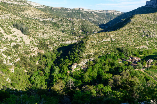 Elevated view of a lush green canyon with scattered stone buildings nestled among dense trees in Navacelles. Sunlight creates dramatic contrasts across the rugged limestone slopes and vibrant valley.