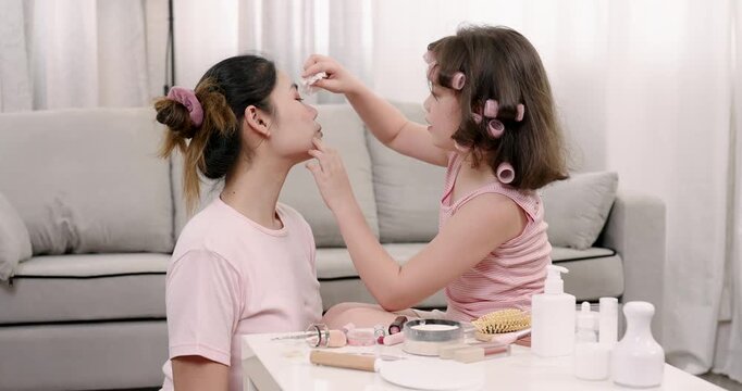 Multiracial girl holding cotton pad gently cleaning Asian woman&rsquo;s cheek during playful makeover session, surrounded by beauty items, expressing tenderness and loving interaction