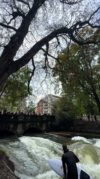 Wet suit surfers River Surfing morning Eisbachwelle Eisbach Isar River Munich Germany vertical tourist attraction street view Autumn Fall Englischer Garten standing water action sports stunts pan down