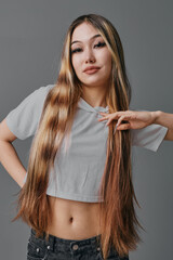 Young woman with long straight hair posing in casual cropped t shirt and jeans on gray background. Beauty model with natural makeup and confident expression looking at camera.