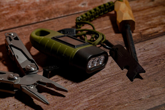 Survival gear kit, including a multitool, a solar flashlight, and a fire starter, arranged on a rustic wooden surface