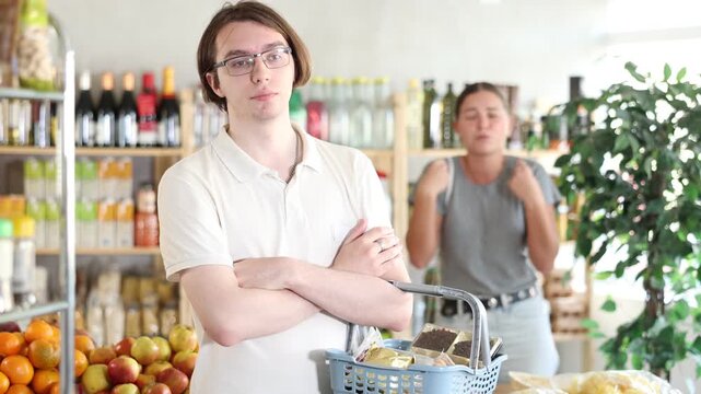 In shop girl stands behind husband, hurling abuse and indignation at him. In trading floor, sullen man stands with shop basket, swears with wife and listens to womans accusations. 
