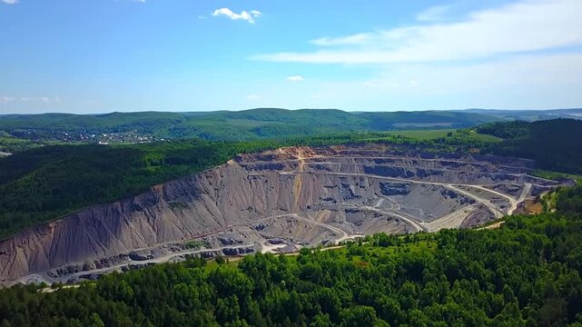 Aerial view depicts an expansive quarry etched into a hillside surrounded by lush forests under a bright blue sky