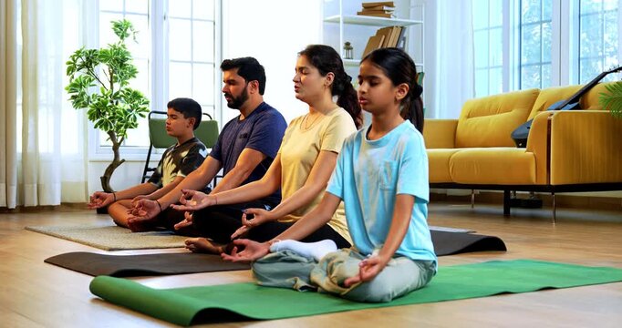Indian young family doing omkar meditation pranayama at home chanting aum breathing yoga together indoors during peaceful morning hours in a modern living room promoting yogic lifestyle