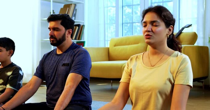Indian young family doing omkar meditation pranayama at home chanting aum breathing yoga together indoors during peaceful morning hours in a modern living room promoting yogic lifestyle