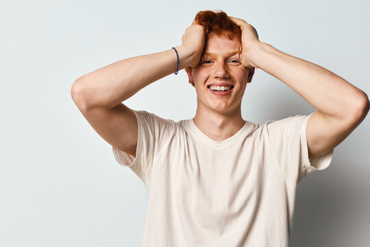 Young man with red hair smiling happily and holding his head with both hands in casual white t shirt against plain background.