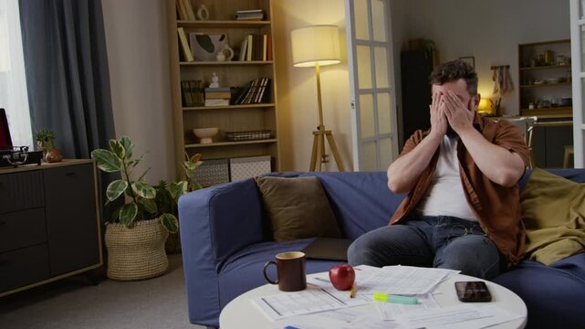 Medium shot of exhausted adult man closing laptop while sitting on couch and working from home, various documents scattered on table, copy space