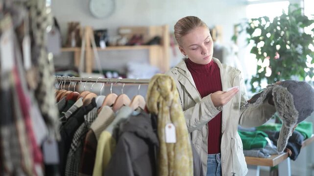 Attentive teen girl customer holding warm fur hat during winter sale in retail outlet 