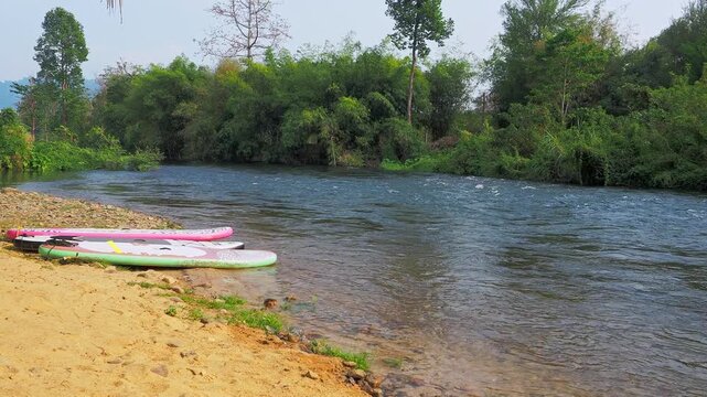 Paddleboards rest on the sandy bank of Khlong Wang Takhrai, ready for an adventure along the river's forested path.