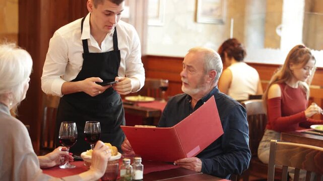 Polite waiter taking order from elderly couple during relaxed dinner in comfortable restaurant setting. High quality 4k footage