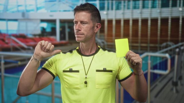 Man referee holding yellow card and thumb to chest, wearing neon shirt and whistle on cruise deck; warning penalty discipline.