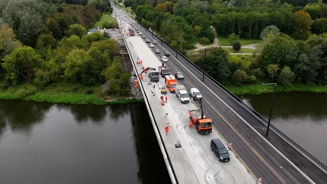 Drone aerial of bridge reconstruction over a river with roadworks crew, orange trucks, and lane closure as cars pass, showing transport infrastructure maintenance in daytime.