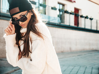 Obraz premium Portrait of beautiful smiling model. Female dressed in warm hipster white sweater and beanie. Trendy woman posing in the street at sunset in sunglasses