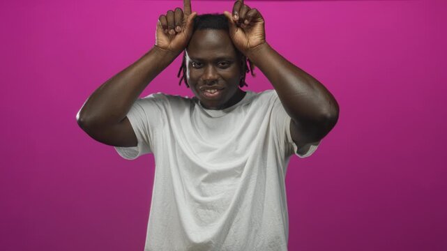 Young african american man with hands making horn gesture in studio pink backdrop wearing white t shirt and dreadlocks; playful mischief confidence.