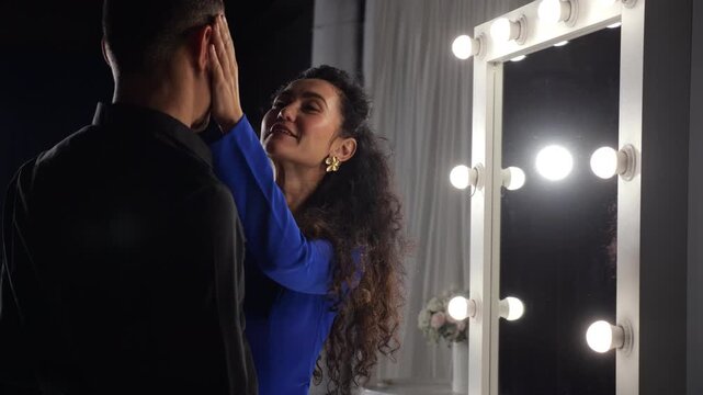 Dressing Room Mirror Woman Touches Partner Face Under Spotlight, Playful Flirtation, Dramatic Bulbs, Blue Dress, Warm Smiles, Gentle Caress, Intimate Backstage Atmosphere