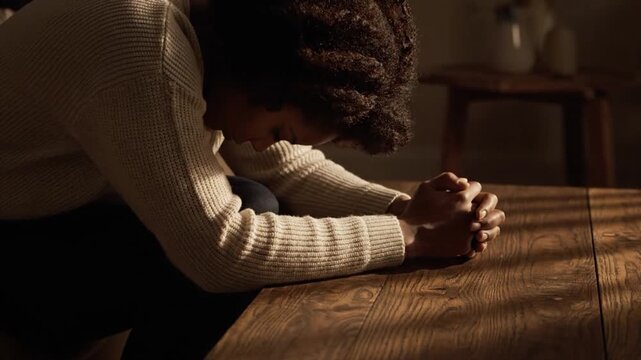 Young African American Woman Sitting at Wooden Table with Head Bowed and Clasped Hands in Prayer or Deep Contemplation in Soft Dramatic Sunlight.