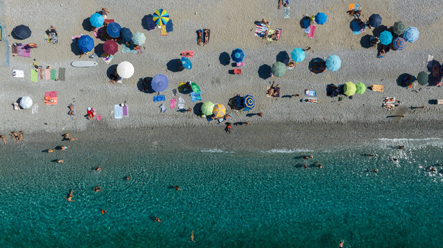 High-angle aerial view of a crowded pebble beach with scattered colorful sun umbrellas and people swimming in crystal-clear turquoise water. Zenithal shot of an italian coast. It is a sunny summer day