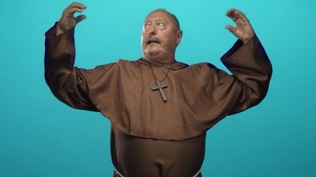 Brown robed man with cross necklace raises hands in studio, looking startled with open mouth and upward gaze while gesturing dramatically; surprise devotion.