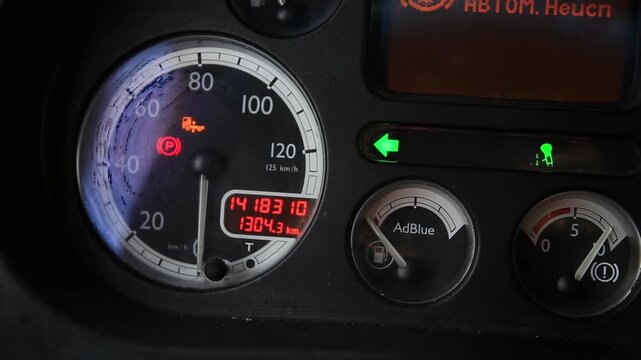 Truck dashboard close up. Speedometer at zero. Odometer shows mileage. Parking brake engaged, engine warning. Turn signal active. AdBlue gauge. Vehicle information display.