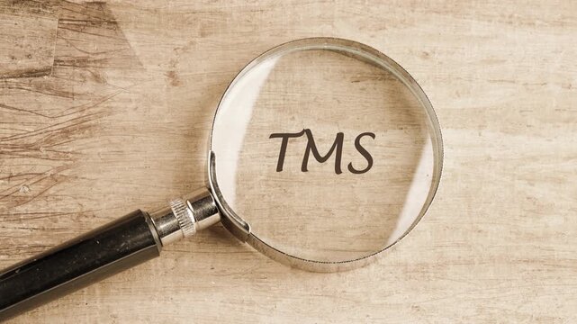 The letters TMS Transcranial Magnetic Stimulation become clearly visible as a magnifying glass is carefully positioned over a textured light wooden table