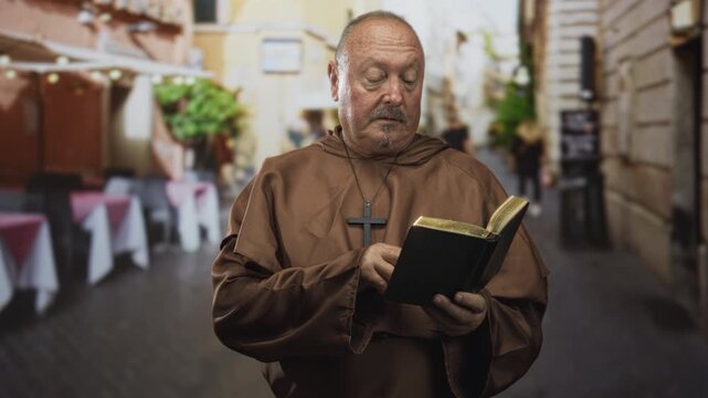 Man monk holding book with hands, reading and looking upward on a street cafe building while wearing robe and cross pendant; contemplation prayer serenity.