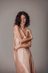 Fototapeta premium The model is a young beautiful brunette girl with curls on a gray background in a pink silk dress and glasses