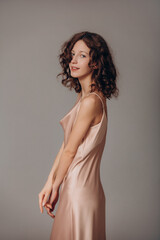 Obraz premium The model is a young beautiful brunette girl with curls on a gray background in a pink silk dress