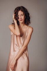 Fototapeta premium The model is a young beautiful brunette girl with curls on a gray background in a pink silk dress