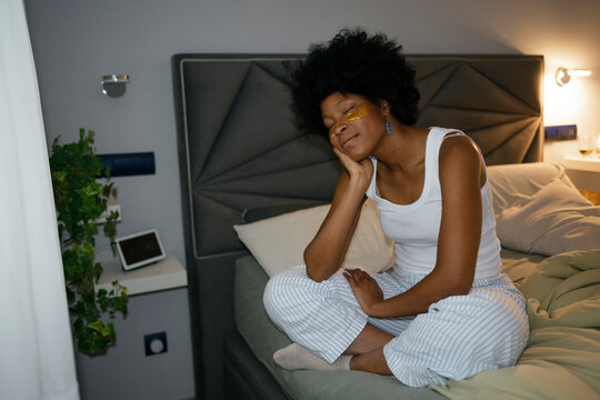 Thoughtful woman with eye patches sitting on bed in evening bedroom