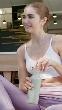 Vertical video: Smiling woman in bra lilac leggings twisting cap in cafe, sipping bottle to hydrate