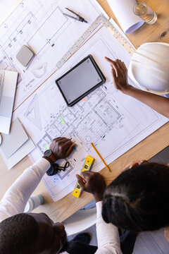 Diverse coworkers examining blueprints at drafting table with digital tablet, spirit level