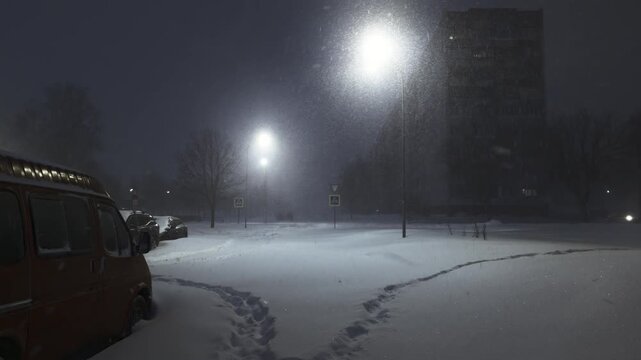 Urban street scene at night during heavy snowstorm illuminating falling snowflakes, ground covered by deep snow, parked van, distant building and footprints leading away