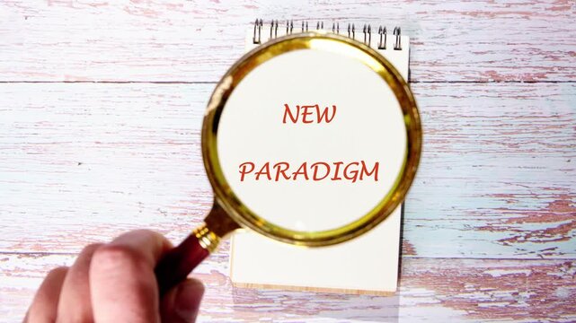 A hand uses a magnifying glass to bring the words NEW PARADIGM into focus. The text is written on a white notepad, resting on a rustic light wood table