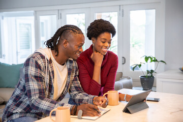 Fototapeta premium African american couple reviewing tablet and writing in notebook at kitchen countertop