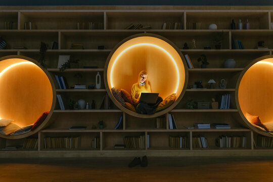 Person relaxing with laptop in cozy, illuminated library coworking space