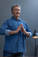 Senior man standing against grey wall in denim shirt making steeple gesture near black lamp