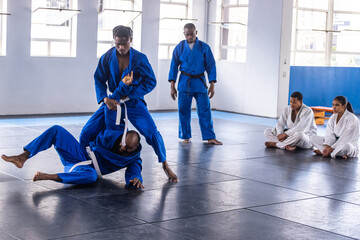 Diverse martial arts class grappling, throwing in blue gis at dojo on mats, wooden boards