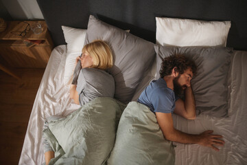 In a modern bedroom, a couple lies in bed facing away from each other, wrapped in blankets. The dim lighting creates a sense of separation and tension between them late at night. © Stockphotodirectors