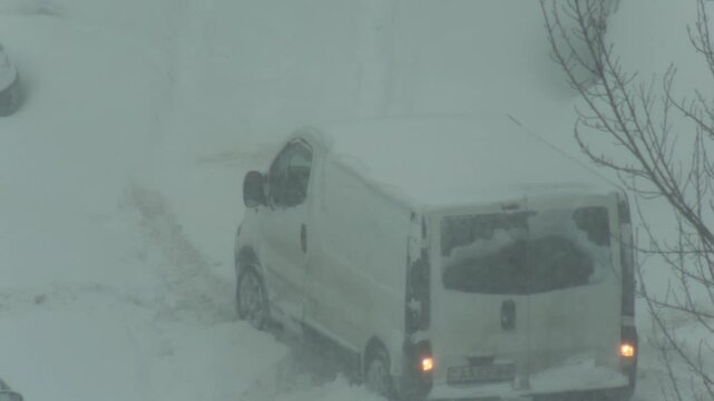 Delivery van immobilized by deep snow accumulation on unplowed road surface. Vehicle rear view, wheels spinning, attempting to move in blizzard conditions.