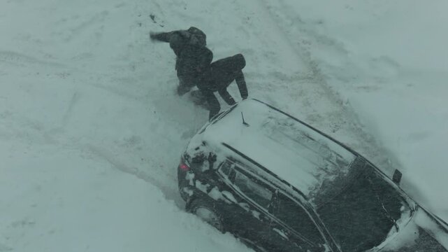 Overhead view people pushing passenger car trapped deep snowdrift during intense winter blizzard. Heavy snowfall covers ground, creating difficult travel conditions. Vehicle recovery effort.