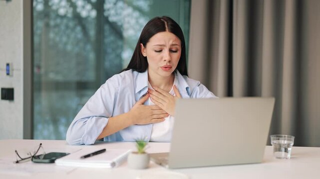 Woman experiencing sudden chest pain and breathing difficulties at desk. Expresses severe discomfort, anxiety, and fear, suggesting urgent medical concern.