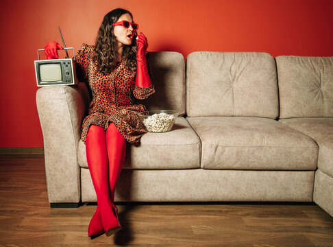 Stylish person in leopard dress eating popcorn on sofa with retro TV