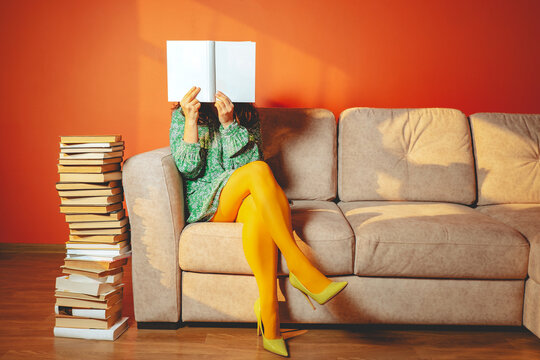 Person in yellow tights reading on sofa with stack of books in living room