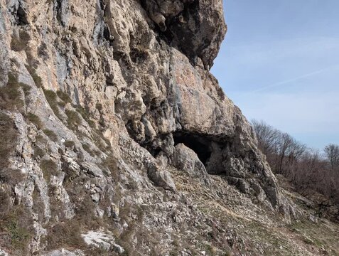Una cavit&agrave; naturale si apre al centro di una parete rocciosa, incastonata in un pendio aspro con vegetazione rada.