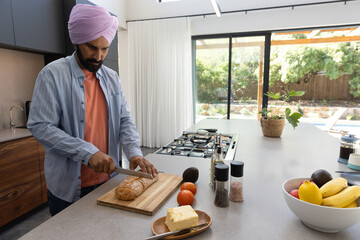 Mid adult indian man slicing bread, tomatoes, avocado on board at kitchen island, copy space © wavebreak3