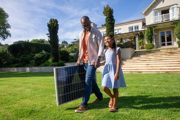 Obraz premium Diverse father and daughter walking across lush lawn at suburban home, carrying solar panel