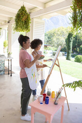 Obraz premium Diverse mother and daughter painting on wooden easel on veranda in aprons amid hanging plants